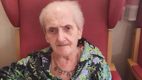 Dudley Metropolitan Borough Council Rita Barnsley has short grey hair and is wearing a blouse. She sits on a large red chair. She is also wearing a necklace.
