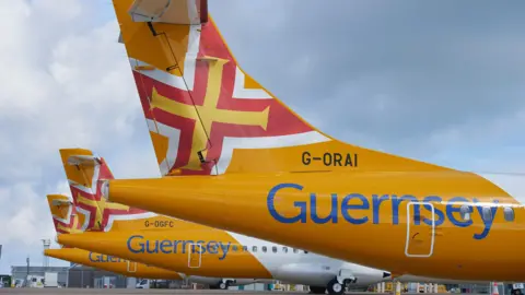Three Aurigny planes are parked on tarmac. Two of their numbers can be seen: G-ORAI and G-OGFC and all three have Guernsey printed in blue lettering over the yellow bodies. The tail fins feature the Guernsey flag which is a yellow cross inside a red cross which is outlined in white.