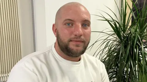 Phil Bodmer/BBC Andrew Dunn has a shaved head and short brown beard and is wearing a white jumper