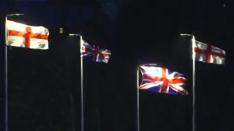 Four flags flying from lamp-posts in Exeter - a St George's cross on the left and right and two Union flags in the centre.
