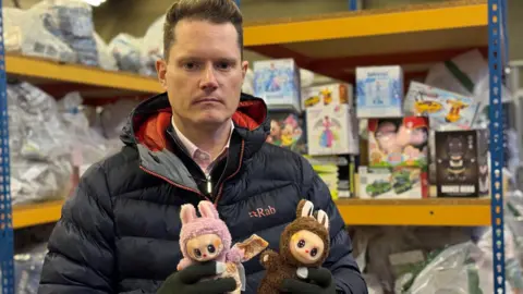 Rhys Hurley, Head of Intelligence Partnerships at the IPO holds up two fake Labubu dolls, the one on the left is pink and the one on the right is brown. He is in a warehouse full of seized goods. Behind him you can see other toys on shelves that have been seized.