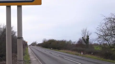 A rural two-lane road stretches into the distance under a cloudy sky, bordered by grassy fields and leafless trees. A red car is partially visible on the right side, moving along the road.
