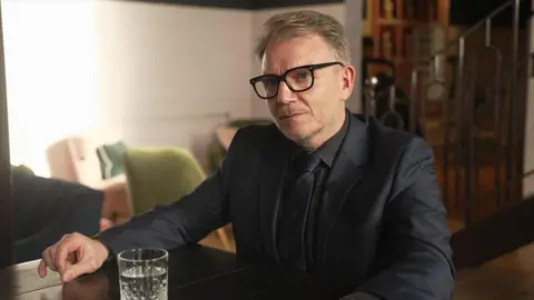 Ian Palmer/BBC A middle-aged man with glasses wearing a black suite, shirt and tie sat in a room 