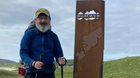 Charles Bain Smith stands beside the Hebridean Way sign in outdor walking gear and holding walking poles, with the Scottish coastline and mountains in the background.