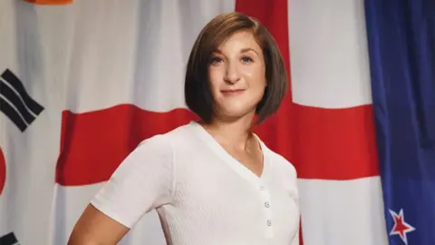 A headshot of Robyn Cowen, wearing a white top, smiling, with an England flag behind her.