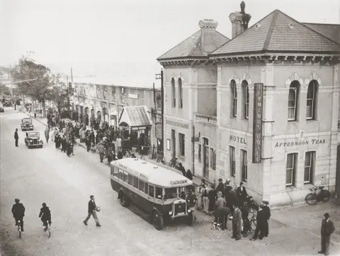 Societe Jersiaise Photographic Archive (008287) A black and white image of a bus on a busy early 20th Century street, with shops and a hotel behind it.