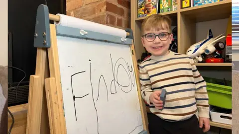 Finley is standing next to a whiteboard wearing a stripy top and glasses. 