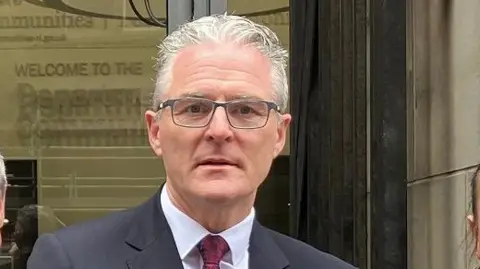 Jarlath Burns has grey short hair and wears glasses with a grey frame.
He is wearing a grey suit, white shirt and burgundy tie.