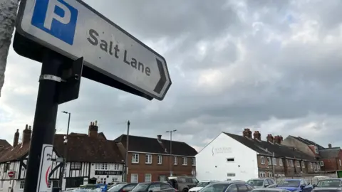 A pointed road sign directed at a car park under a cloudy sky. The sign says "Salt Lane". The tops of cars can be seen and terraced buildings are in the background.
