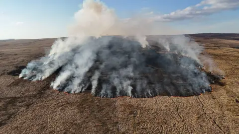 An aerial picture of the large mountain fire actively burning. Lots of grey smoke is rising into the sky. 