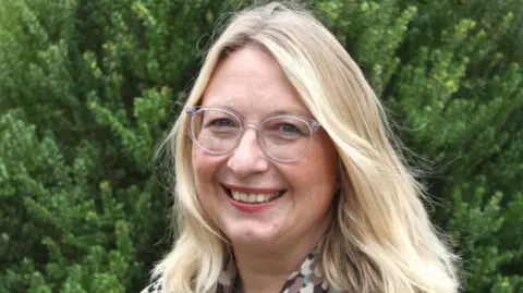 St Elizabeth Hospice Judi Newman has long blonde hair and is wearing clear framed rounded glasses. She is smiling, and standing near a fir hedge. Part of her blouse, white, patterend with beige, blue and brown shapes, is partly visible.