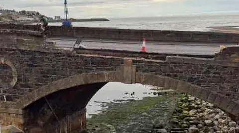 Fubar News Collapsed section of Banff Bridge. A section of brickwork has falled to the area below. The sea is in the backgroundm with a single orange and white cone on the bridge. 