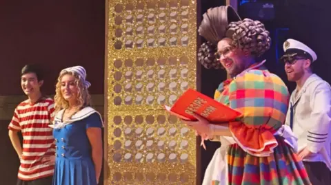 Juliette Thymi James Wood wearing a multi-coloured dress and a large brown wig. He is holding a large book and standing on a stage with three other performers. 