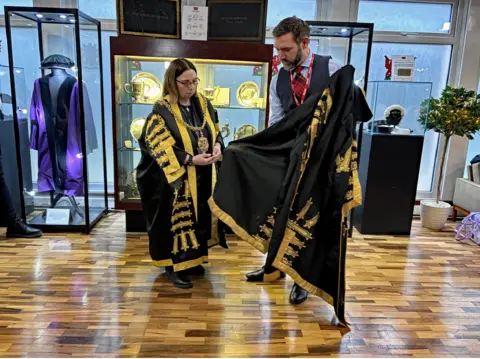 Ian Duncan Lee Paton shows Jeanette Whalen the old robe. It is very similar to the one she is wearing. It is black and has gold trim. The mayor is also wearing her large gold chain around her neck