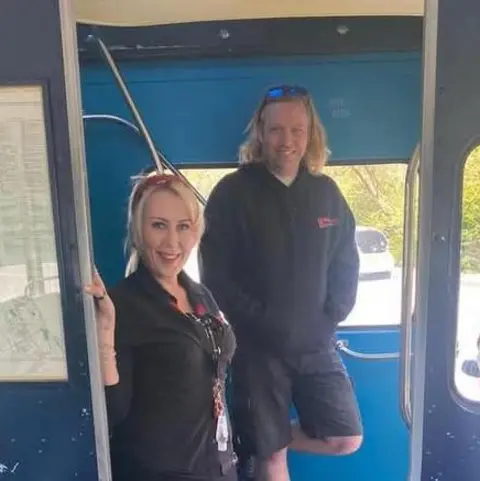 Karen Hall Karen and Kev stand inside a blue bus. They both have black shirts on and Kev has black shorts. They both smile at the camera and they both have blonde hair.