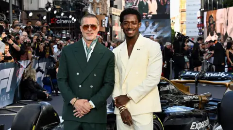 Getty Images Brad Pitt and Damson Idris attend the European Premiere of F1 The Movie at Cineworld, Leicester Square on June 23, 2025 in London. Pitt, left, wears aviator sunglasses and a dark green double-breasted suit with a white shirt and green cravat. Idris wears a cream suit over a bare chest with a gold necklace with green gem stones. They pose in front of a F1 racing car, surrounded by crowds at the premiere.