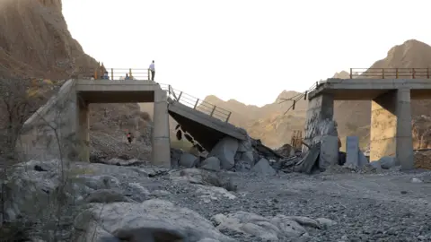 A damaged bridge with people standing on the remaining structure with mountains in the background, and rubble and rocks in the foreground.