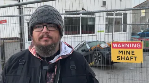 A man with a beard and a black beanie hat looks at the camera - behind him is his car in the mineshaft