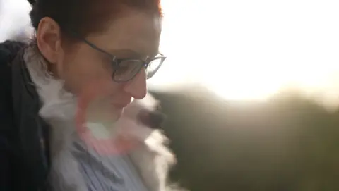 A close-up of Leanne, a woman with red hair and dark-framed glasses, looking down at the ground. The image is shot against bright sunlight shining in the out-of-focus background.
