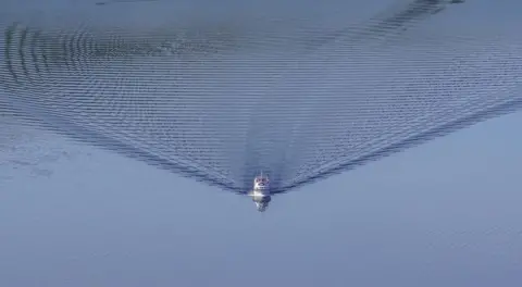 Frank Urban A boat sails across the calm blue water creating ripples behind it 