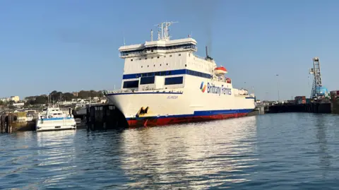 BBC A Brittany Ferries vessel called Islander docked in Guernsey. The ferry is mostly white with some blue stripes. It is a sunny day.