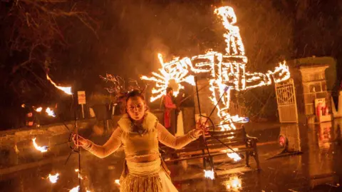 Kildare County Council/Allen Kiely A woman dressed in festival costume stands in front of a large, burning St Brigid's cross, illuminating the night sky as part of festival celebrations in Maynooth