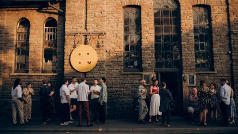 A crowd of people stand outside a converted church building which has tall windows and light coloured stonework.