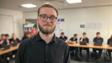 A blonde bearded man wearing a black shirt and glasses smiling at the camera. A row of students are sitting at desks with laptops behind him.