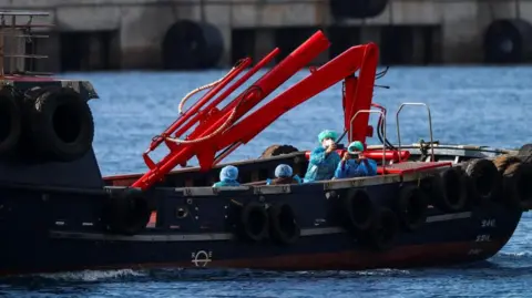 Passengers wearing blue plastic overcoats, green head coverings and face masks hold up their phones as they travel on a boat from a cruise ship to the shore in Tenerife.