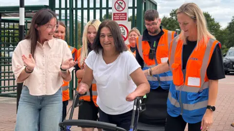 A woman wearing a white t-shirt is seen standing with the help of a stroller on wheels. She is surrounded by people who are cheering her on, as she uses a pelican crossing.