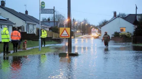 People walking through floodwater near homes. Several people are wearing high vis jackets and one is walking a dog.