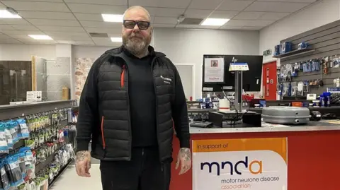 John Fairhall/BBC Michael King stands in a room as he smiles at the camera. He has short grey hair and is wearing glasses as well as a black puffer coat. 