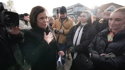 PA Media Mary Lou McDonald with shoulder length dark hair holds her hand to her chest as she talks to two female students both wearing dark coats at an outdoor event in Dublin