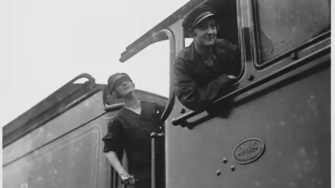 A black and white image of two women driving a steam train. They are both wearing overalls as well as caps. One is in the driver's seat and the other just behind.