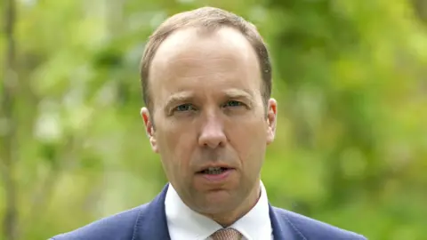 A man standing outside. He has thin light brown hair and is frowning slightly with his mouth open. He is wearing a blue suit with a white shirt and a light brown tie.