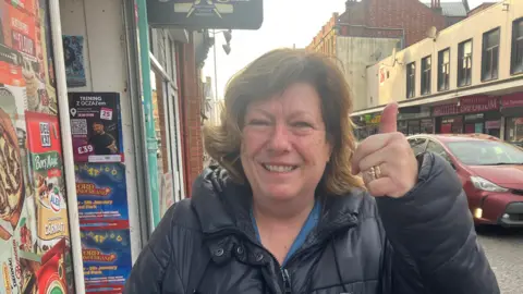 Liz smiles and gives a thumbs up to the camera as she stands on a pavement with a red car driving down the street behind her. She has brown hair and is wearing a black puffer jacket.  