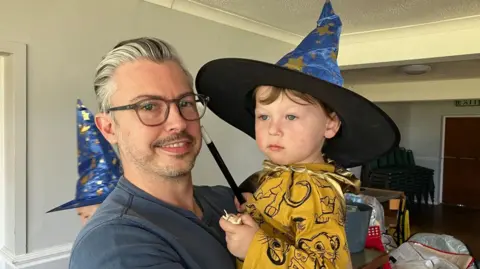 A man with grey-streaked hair and glasses holding a small child in a gold top and a wizards hat