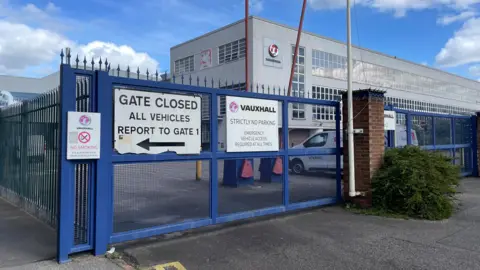 The closed blue gates outside the Vauxhall plant. A grey building with many windows and a Vauxhall logo is behind the gates.