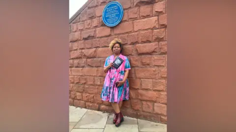 Michelle Adamson/BBC Karen Gabay in a large multi-coloured skirt stands below the blue plaque