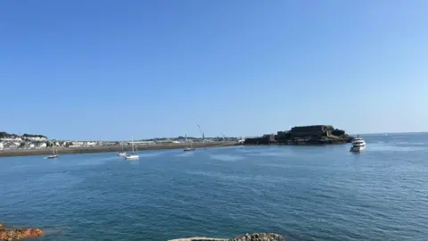 Havelet Bay - A picture of the blue sea, with a few sail small boats with their anchor dropped to the left aqnd a large grey yacht to the right. In the background Castle Cornet is pictured, a granite fortress, with the Castle Emplacement rolling down the left. 