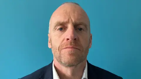 James is looking at the camera with a plain expression. He is wearing a suit jacket and white shirt in front of a blue background.