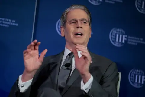 Scott Bessent raises his hands while talking, sitting in front of a dark blue screen.