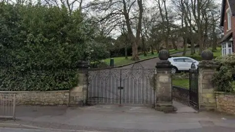 Black metal gates across a paved path leads up between trees off a road.