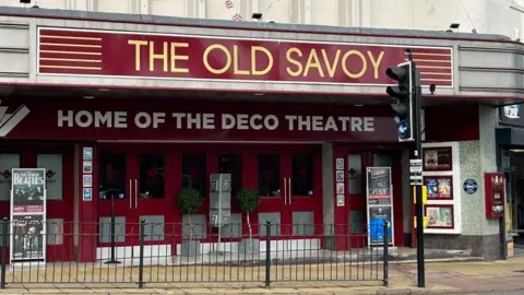 Ollie Conopo/BBC Red facade of theater building with six high windows. Posters advertising events are visible on both sides. The legend above the doors says "The Old Savoy - Home of the Deco Theatre". There is a black railing and a traffic light outside the theater.