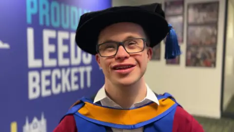 George Webster, who has glasses and is smiling, wearing his graduation robes which are red and blue, he is standing in front of some Leeds Beckett branding and pictures.