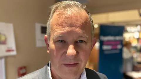 Luke Deal/BBC Andy Mellen smiles at the camera. He has short grey hair. He wears a grey suit with a white shirt underneath.