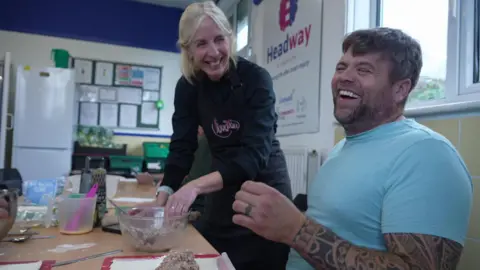 BBC A man wearing a blue T-shirt laughing and smiling with a lady with blonde hair. The man has tattoos up his arm. The woman is wearing a black apron.