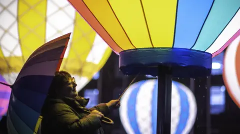 Andre Pattenden A woman with a colourful umbrella appears to be holding a stick as she admires an illuminated display of hot air balloons, also in rainbow colours. She is wearing large glasses and a raincoat, and a few drops of rain can be seen.