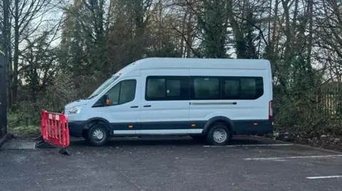 A white minibus is parked in a car park with a red plastic fence placed in front of it. The car park is surrounded by a mental fence with vegetation woven around it.
