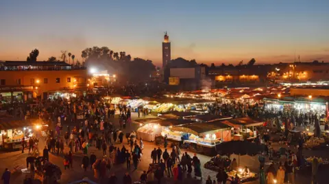 Jeff Overs/BBC Una vista general de la noche de Jamaa El Fnna en Marrakech. Las luces naranjas brillan desde el otro lado de la plaza y se puede ver a las personas caminando por los puestos del mercado. 